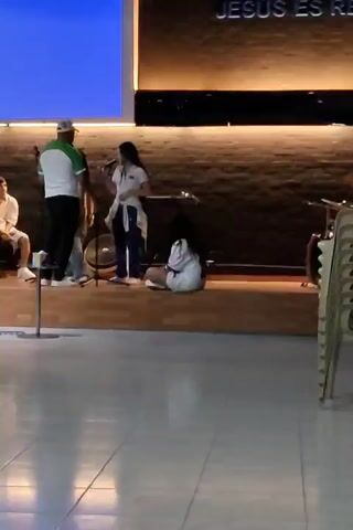 Here is a concise description of the video:

The video shows a dimly lit stage with a band preparing for a performance. A young woman stands center stage, singing into a microphone. She is dressed in a white shirt, a white sweater tied around her waist, and blue pants. Another young woman sits on the stage to her right. To the left of the singer is a man dressed in a white shirt with green accents, black pants, and a hat. Behind them, a textured wall serves as a backdrop. There is a keyboard on the stage, along with percussion instruments. Above the singer, there is a screen displaying a blue color. The stage is wooden, and the floor in front of the stage appears to be polished tile.
