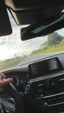 A first-person point-of-view video from inside a car driving on a highway. The driver's left hand is on the steering wheel, which is black. The dashboard is visible, showing a speedometer, other gauges, and a digital display screen. The screen shows a map with a route highlighted in blue and some icons on the left side, possibly for navigation or other car functions. The road ahead is clear, with a few cars in the distance. Trees and green vegetation line the sides of the highway. The sky is partly cloudy with a bright horizon, suggesting either early morning or late afternoon. A billboard is visible on the right side of the road.
