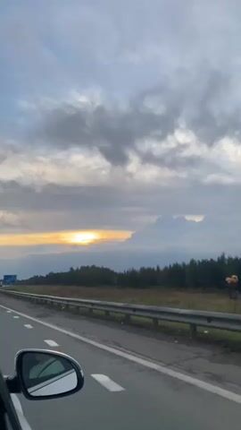 The video shows a view from inside a moving car on a highway. The sun is setting, casting a warm glow on the horizon. The sky is filled with clouds, some of which are dark and heavy, while others are lighter and wispy. A line of trees runs along the right side of the highway, and a guardrail separates the road from the trees. A blue sign with white text is visible on the right side of the highway, indicating distances to various cities. The car's side mirror is visible in the lower left corner of the frame.