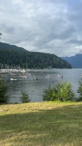 A wide shot shows a body of water with numerous people on paddleboards and kayaks. In the background, a forested hillside rises steeply, with a marina full of sailboats to the left. The sky is cloudy. The foreground consists of a grassy embankment. The people on the water appear to be engaged in a group activity or race.