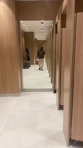 The video shows a person standing in a hallway lined with changing rooms. The hallway has a light-colored tiled floor and light wood-paneled walls. Overhead, a series of recessed lights illuminate the space. The person, wearing a dark short-sleeved shirt, light-colored pants, and sneakers, is holding a smartphone and taking a selfie in a large mirror that spans the width of the hallway.

In the reflection, the person is walking towards the mirror. They are carrying a small, orange shopping bag in their left hand. To the left of the person in the reflection, there is a rack with several garments hanging on it and a metal cart. The person then raises their phone higher, seemingly to capture more of the ceiling or the lights above. The activity appears to be a person taking a selfie in a retail environment, likely a store with changing rooms.

The setting suggests an indoor location, possibly a department store or a boutique. The lighting indicates it is daytime or that the store is well-lit. There are no visible weather cues. The person's expression is neutral as they focus on taking the photo. No text is visible in the video. The location context of Chennai, India, is provided, but there are no specific visual cues within the frame to confirm this location.