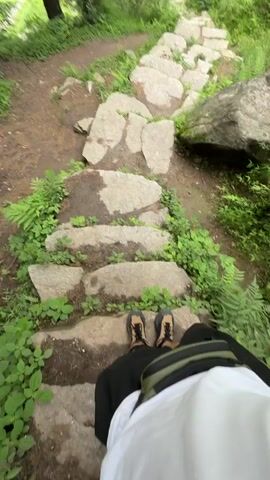 The video shows a point-of-view shot of someone walking up a stone pathway through a green, overgrown environment. The pathway is made of irregularly shaped, flat stones laid out in a staggered pattern. Lush green ferns and other foliage grow on either side of the path and in the background. The person walking is wearing black shorts and sandals. A green and black bag is visible on their back, and a white garment is also visible. The camera movement is shaky, indicating the person is actively walking. The overall impression is of a hike or walk in a natural, possibly mountainous or forested area.