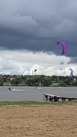 A kitesurfer is seen from a sandy beach as they ride on a lake. The kitesurfer is on the left side of the frame, moving from left to right. A purple kite is visible in the upper right quadrant of the frame. Another smaller blue kite is visible in the distance. A wooden pier extends into the water on the right side of the frame. Trees and buildings are visible in the background. The sky is cloudy and dark.