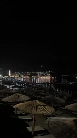 This media file captures a nighttime view of a beach in Boreti, Montenegro. The scene is dominated by the dark sky, with the foreground occupied by numerous beach umbrellas and lounge chairs, many appearing to be made of natural materials like straw. The umbrellas are closed and set up in neat rows on the sandy beach.

In the background, the lights of a coastal town twinkle brightly, reflecting on the calm surface of the water. The lights suggest a lively town or resort area. The overall impression is one of a peaceful and serene nighttime ambiance. The time of day is clearly night, and no specific weather conditions are apparent, though the calm water implies clear skies or at least no significant wind or rain. The location is identifiable as a beach in Boreti, Montenegro, indicated by the presence of beach furniture and the distant town lights along the coast.

There are no people visible in the frame, and the focus is on the landscape and the man-made beach amenities. The emotion evoked is one of tranquility and quiet beauty, showcasing the allure of the coastline after sunset. No text is visible in the media file.
