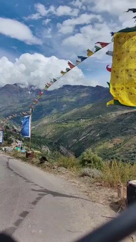 The video shows a roadside view of a mountain village. The road curves to the left, with a steep drop on the right overlooking a valley. Several prayer flags are strung across the foreground and middle ground, fluttering in the wind. A tall blue and white flag is also visible on a pole. The village itself is nestled on the mountainside, with buildings clustered together and green terraced fields in the valley. The mountains in the background are rugged and green, with some snow-capped peaks visible under a bright blue sky with fluffy white clouds.