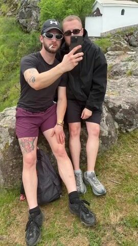 Two men are taking a selfie while smoking cigarettes. They are outdoors, seated on a large rock in front of a hill covered in vegetation. At the top of the hill, there is a small white building. The man on the left wears a black cap, a black t-shirt, sunglasses, and burgundy shorts. He has tattoos on his arms and leg. The man on the right wears sunglasses, a black hoodie, and black shorts. The sky is overcast. A black backpack sits on the ground next to the men. Someone in the background says "Wow! Wow!".