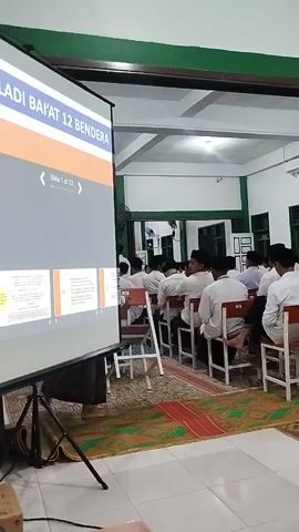 A group of young men, mostly wearing white shirts and black caps, are seated in chairs facing a projector screen in what appears to be a classroom or meeting room. The screen displays a presentation with the title "ADI BAI'AT 12 BENDERA" and "Slide 1 of 22." The room has green accents on the walls and ceiling. A fan is mounted on the wall. The floor is covered with a patterned rug. Some individuals are looking at the screen, while others appear to be looking elsewhere or interacting with the person next to them. One person in the foreground is partially visible, seemingly operating the presentation equipment. The lighting is artificial, suggesting an indoor setting. The overall atmosphere suggests an educational or informational session.