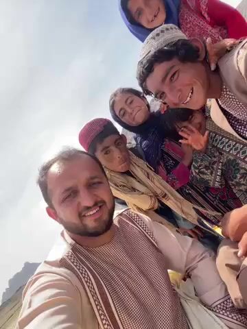 In a bright daylight selfie video, an adult male with a beard and traditional attire is in the foreground, holding the camera. Behind him, a group of five people, including an adult female and four children, are also visible. They appear to be of Afghan ethnicity, wearing traditional clothing including head coverings for the women and children, and patterned shirts/dresses.

The group is joyful and expressive. The adult male is smiling brightly at the camera. The children behind him are laughing, making playful faces, and gesturing with their hands. One young girl smiles widely and puts her hands to her cheeks, while another girl raises her hand in a waving or playful gesture. An older boy with a white cap also smiles and laughs. The adult female behind them is also smiling.

The setting appears to be outdoors in a mountainous region, with a clear blue sky overhead and mountains visible in the background, suggesting a rural environment in Ashoqah, Afghanistan. The overall mood is cheerful and friendly. No visible text is present in the video.