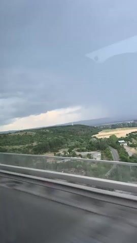 The video shows a view from a moving vehicle, looking out over a landscape. The foreground consists of the vehicle's interior, including a blurry gray surface and a section of a glass barrier with metal trim, suggesting it might be a train or bus. Beyond the barrier, the landscape is visible. It features rolling green hills covered in trees and vegetation. A road winds through the terrain, leading towards some fields in the distance. The sky is overcast with dark, heavy clouds, hinting at potential rain or a storm. A tall, white tower-like structure is visible on one of the hills. The lighting is subdued due to the cloudy weather.