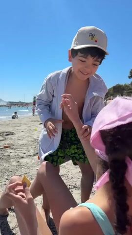 On a sunny beach, a young boy wearing a light grey robe and a beige baseball cap smiles at a girl sitting on the sand. The girl wears a light blue top and a pink paisley bandana. The boy takes something from the girl’s hand, then looks towards the ocean. The beach has multiple people in the background, and the water is a clear blue. There is an orange ball on the sand near the boy.