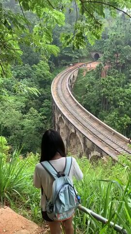 The video opens with a high-angle, medium shot of a curved railway bridge made of stone arches, spanning across a lush, green valley. Dense foliage with large leaves frames the top of the shot. In the foreground, the back of a young woman wearing a light-colored t-shirt and a light blue backpack is visible. She is standing with her back to the camera, looking out at the bridge. The railway tracks are clearly visible on the bridge, which curves around to the left. In the distance, a tunnel entrance can be seen on the hillside. Several small figures, presumably people, are standing on the ground near the tunnel entrance and along the railway line. The environment is characterized by extensive greenery, including tall trees and undergrowth, suggesting a tropical or sub-tropical setting. The lighting appears to be overcast or diffused, indicating a possibly cloudy day or early morning/late afternoon light. The video is static for the duration of the clip.