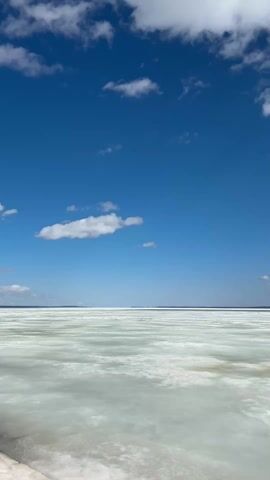 The camera faces directly toward a seemingly frozen lake, the ice appearing fractured and partially melted. Above, a clear blue sky is punctuated by scattered white clouds, some large and fluffy, others smaller and more wispy. A thin dark line is visible on the horizon, possibly indicating the distant shoreline or treeline. No people, objects, or explicit actions are visible in the video frame.