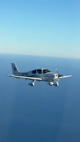 The video shows an aerial view of a white Cirrus SR22 aircraft with the registration number N219EL flying over a body of water. The sky is clear and blue. The aircraft is viewed from the side and slightly from above, with its propeller spinning. The fuselage of the aircraft has "LA" printed on the tail fin. The video appears to be a screen recording or a video taken from another aircraft.