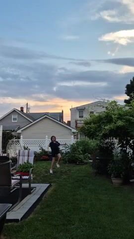 A person wearing a dark t-shirt and dark shorts walks from left to right across a grassy backyard. The person appears to be holding something in their hands. The backyard contains several houses and fences in the background. The sky above is cloudy with hints of orange and yellow from a sunset.
