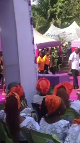 The video shows a gathering of people outdoors under tents labeled "Goshencity.com." The area is decorated with purple and white fabric. Many of the attendees are wearing matching orange headscarves and white lace outfits. On a raised stage with a purple carpet, several people are walking and interacting. Some of them are wearing bright yellow shirts, while others are in traditional African clothing. One person is carrying a tray of objects, possibly food or gifts. The scene appears to be a celebratory event or ceremony.