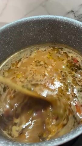 A close-up overhead shot shows a wooden spoon stirring a bubbling liquid in a gray saucepan on a black stovetop. The liquid is a murky brown with visible pieces of red, green, and white ingredients. The stovetop is dark and appears to be a flat electric surface. The background is a blurry marble-patterned backsplash. The lighting is soft and natural. The video is short and shows the liquid being stirred.