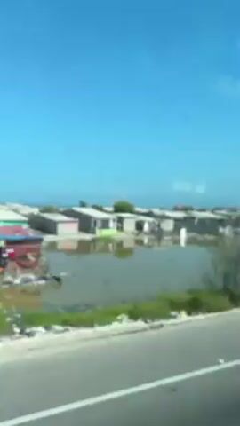 The video shows a view of a settlement next to a body of water, possibly a lagoon or river, from a moving vehicle. The buildings appear to be simple, possibly residential structures, and are situated near the water's edge. The landscape around the buildings is mostly barren, with patches of vegetation and debris. The sky is clear and blue. The video is filmed through a window, and there are some reflections and imperfections on the glass. The focus is not sharp, and the details are somewhat blurred due to the movement of the vehicle.