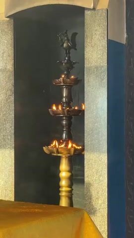 The video shows a tall, dark, multi-tiered oil lamp with flames burning on each tier. The lamp has a gold colored base and a figure of a bird perched at the top. The lamp is inside an alcove, with stone walls and a gold colored cloth draped in front of it.