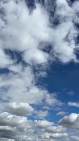 This is a vertical, full-screen video of a blue sky with scattered white clouds. The camera is tilted slightly upward and appears to be moving slowly, or the clouds are drifting, as they subtly shift in position within the frame. The clouds vary in size and density, with some appearing wispy and thin, while others are thicker and more substantial, casting soft shadows. The lighting suggests it is daytime, possibly with the sun behind the viewer or slightly off-camera, illuminating the clouds from above. There are no people, objects, or any other environmental elements visible in the frame besides the sky and clouds.
