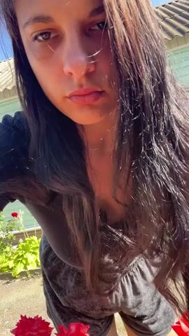 The video shows a young woman with long, dark hair wearing a black t-shirt and dark shorts. She is outdoors in what appears to be a garden. She is leaning down and looking at a bush of red roses. The background shows a house with a green siding and a fence. The lighting suggests it is daytime and sunny. The woman's expression is serious and she is not smiling. The camera angle is close up, focusing on the woman's face and the roses.