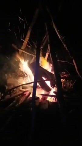 A campfire burns brightly in the dark, with embers and sparks flying upwards. Several logs are stacked in a teepee formation, and flames are consuming the wood. The surrounding area is dark and indistinct.