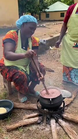 A woman wearing a patterned top, apron, patterned pants, and blue hairnet kneels on the ground and stirs a pot of thick, reddish-brown substance with a long wooden spoon. The pot sits on a stand over a wood fire. Next to her, another person in a green apron and patterned skirt stands partially visible. In the background are a few buildings and trees, with a black bucket containing something floating in water to the left. Smoke rises from the fire, and the woman smiles as she works. The ground is dirt and scattered leaves.