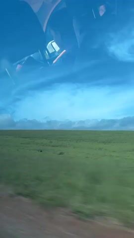 The video shows a view from inside a vehicle traveling along a dirt road next to a large green field under a cloudy blue sky. The reflection of the vehicle's interior, including what appears to be a person driving, is visible on the windshield. The scenery is blurred due to motion, creating a sense of speed. A tree is seen passing by on the right side of the frame.