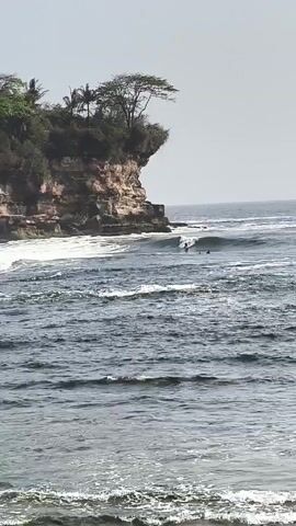 The video shows a surfer riding a wave near a cliff. There are other surfers in the water further out. The water is dark and choppy with white foam. The cliff is tall and rocky with trees on top. The sky is clear and bright.