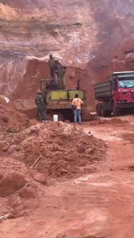 A slow-motion video shows a construction scene. In the foreground, mounds of red dirt are visible, with a few small branches scattered on the ground. In the middle ground, a yellow excavator is positioned, with its arm raised and a bucket extended. Two individuals are standing near the excavator. One, wearing camouflage clothing, stands to the left of the excavator. The other, wearing an orange shirt and blue jeans, stands to the right. A white bucket is on the ground near the excavator. To the right of the excavator, a red dump truck is parked, with its bed empty. In the background, a large reddish-brown cliff face dominates the scene, with evidence of excavation. A figure in camouflage clothing is standing on top of the dump truck's cab, looking towards the cliff. The overall environment appears to be a quarry or a large-scale construction site with significant earthmoving activity. The video is captured at a low angle, emphasizing the scale of the machinery and the terrain.