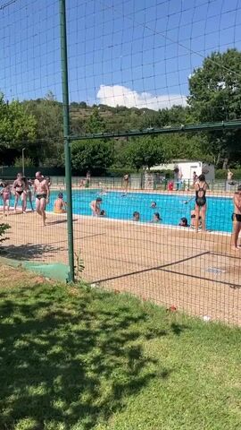The video shows a group of people at an outdoor swimming pool on a sunny day. The pool is a bright blue and filled with water. Several people are in the pool, swimming, splashing, and standing around. Some people are on the pool deck, watching or preparing to enter the water. The pool area is surrounded by a fence, and beyond the fence, there is grass, trees, and a hillside in the distance. The overall scene depicts a recreational activity on a warm day.