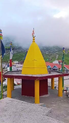 This video captures a tranquil, cloudy mountain landscape. In the foreground, a vibrant yellow, cone-shaped structure stands on a raised platform supported by red and yellow pillars. Prayer flags in various colors flutter from tall poles, adding a splash of color to the scene. Behind the structure, a road winds through a village with buildings featuring red roofs. The backdrop consists of lush green mountains partially obscured by swirling mist, creating a serene and somewhat mystical atmosphere. The overall impression is one of peace and natural beauty.