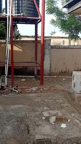 The video shows an outdoor space with two large black water tanks supported by a red metal frame. Below the tanks are pipes and scattered debris. In the background, there is a tan wall and a glimpse of a building. Chickens are walking around near the base of the structure. The camera then pans upward to show the full height of the tanks and the metal frame. Finally, the camera pans to show a tree with birds perched on its branches, and a corrugated metal roof in the background.