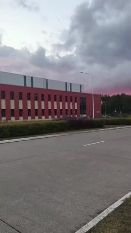 A wide shot shows a road in front of a modern red and gray building. The sky is filled with dramatic, dark clouds that are illuminated by a vibrant pink and orange sunset. There are streetlights and trees visible in the background. Several cars are driving on the road and parked in a lot. A blue and white parking sign with a car icon is visible on the right side of the road. Another traffic sign with a diagram of arrows is also on the left. The camera is stationary.