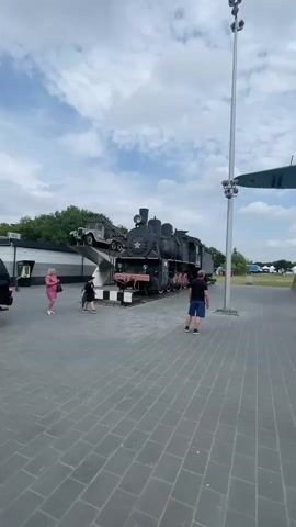 This is a vertical, eye-level, outdoor video taken on a cloudy day. The scene is a museum or outdoor exhibition featuring historical artifacts, likely military in nature. In the foreground, a paved area of gray rectangular tiles stretches towards the background. 

The primary subjects are several large objects: a black steam locomotive with a red star on its front, and a green airplane mounted on a concrete pedestal that resembles a ramp. The locomotive is positioned on tracks and has a small car-like vehicle on top of it. To the right of the airplane, various military vehicles, possibly tanks, are visible in the distance. There are also a few people in the frame: a woman in a pink dress and a child in a black shirt and shorts in the left midground, and a man in a dark blue shirt and shorts walking away from the camera in the right midground. Another person is visible near the military vehicles in the far right.

The lighting suggests it's daytime, likely late morning or afternoon, with the clouds partially obscuring the sun. The setting appears to be a public space designed for display and viewing. There are no visible signs with text in this frame. The overall mood is that of a casual visit to a historical exhibition on a pleasant, albeit cloudy, day.
