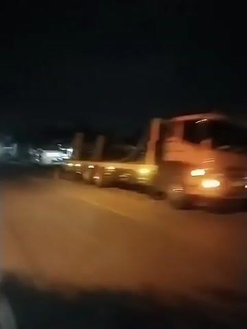 The video shows a road at night.  A large flatbed truck is stopped along the side of the road, with its lights illuminated. Across the street, there is a small building with lights on. A vehicle approaches from the distance, its headlights shining brightly. The road has a white line dividing it into two lanes.  The video is shaky, suggesting it was recorded while moving. The general environment appears to be a residential or semi-rural area.