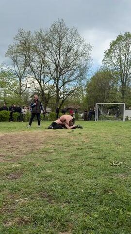 The video shows two men wrestling on a grassy field. One man is shirtless, wearing red shorts, and the other is wearing black. They are grappling on the ground in the center of the frame. A man in a vest stands to the left, appearing to officiate, while a crowd of people watches from the edge of the field. Behind them are trees and buildings. To the right of the wrestlers is a soccer goal. The weather appears overcast.