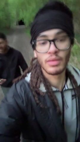 The video depicts three men walking on a dirt path through a forest. The man in the foreground is holding the camera, wearing a black turban, black glasses, and a grey and black jacket. He has long, braided hair. He is talking to the camera. Behind him, two other men walk on the path. The forest is dense with green trees. The lighting is dim.