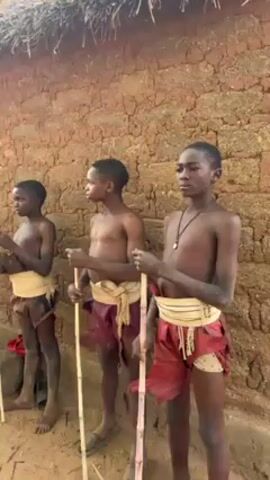 Three shirtless young males stand side-by-side, each holding a thin, light-colored pole. They wear maroon-colored, leaf-like material wrapped around their waists, secured with white fabric and fringe. The boys are looking to the right of the frame. They are standing outside a mud brick structure, with straw on top. Another child is sitting down, left of the three standing.