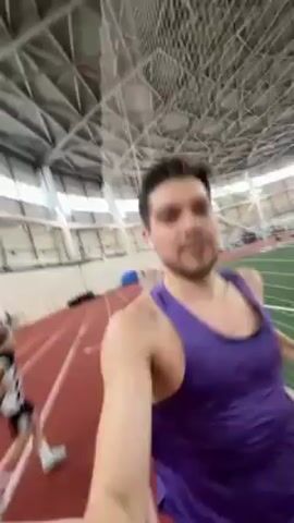 A man wearing a purple tank top jogs along an indoor running track while recording himself on a handheld device. The track is red with white lane markers and curves around a green artificial turf field used for soccer. The man has dark hair, fair skin, and is smiling as he looks at the camera. He appears to be wearing earbuds. Other runners are visible further down the track. The building has a high, arched roof with a complex metal framework and bright overhead lights. The camera angle is slightly tilted and shaky due to the jogging motion.