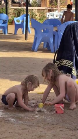 Two young children are squatting on sandy ground, appearing to play in a muddy puddle. The child on the left is wearing dark shorts, while the other child is in white bottoms. They are both touching the mud, possibly shaping or moving it. A small pink bucket sits nearby. In the background, a boy stands on a blue chair, while more empty blue chairs and a towel draped over a stroller are also visible. The setting appears to be a beach or sandy area with outdoor furniture.