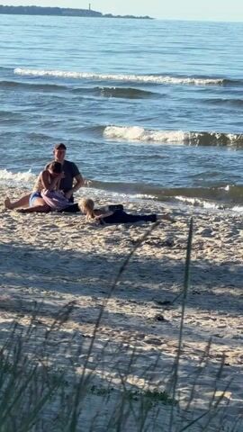 This video shows a sunny day at Plieņciems, Latvia, with a beach and the Baltic Sea as the backdrop. The time of day appears to be late afternoon, with the sun casting a warm glow on the sand and water. Three people are visible on the beach. In the foreground, a person is lying face down on the sand, appearing to stretch or exercise. Further back, a couple is seated on the sand near the water's edge, engaged in conversation. A bottle is also visible near the couple. The overall mood seems relaxed and peaceful, characteristic of a beach outing. The calm sea with gentle waves and clear skies suggest pleasant weather. The presence of dune grass in the foreground adds to the natural beach environment. No visible text is present in the video.