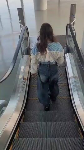 A person walks down an escalator in what appears to be an airport. The person is wearing a light-colored jacket with blue accents and dark, baggy jeans. They have long, dark hair and a small pink bag slung across their body. The escalator has metal railings and the steps are dark with yellow safety lines. The floor at the bottom of the escalator is wet. Large windows overlook an airport tarmac with several airplanes visible in the distance.