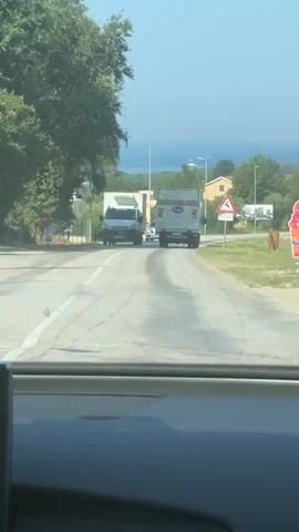 The video shows a view from inside a car driving on a paved road. A white truck is seen on the left side of the road. Further ahead, a white car and a motorcycle are driving in the same direction. A second truck is visible on the right side of the road. On the right side of the road, there is a triangular warning sign with an image of a deer. Trees and foliage are present on both sides of the road. In the distance, the sea and some buildings can be seen under a clear blue sky. The car's dashboard is visible in the foreground.