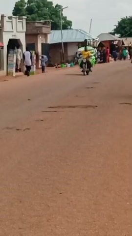 The video shows a dirt road lined with buildings, some appearing to be shops or market stalls. Several people are visible, including individuals walking and riding motorcycles and bicycles. One person is on a motorcycle carrying large green bags. Another person is on a bicycle. There are also some parked motorcycles. The environment appears to be an outdoor setting with trees and a cloudy sky. The road has visible potholes.