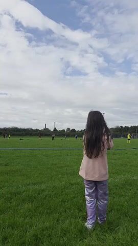A young girl with long dark hair stands on a grassy field, facing away from the camera. She is wearing a light pink shirt and shiny lavender pants. In the distance, several people, likely children, are playing a game of soccer on a marked field. The sky above is cloudy with patches of blue. A person's arm briefly enters the frame from the right, touching the girl's shoulder. The girl then turns slightly and walks forward, holding a dark object in her right hand.