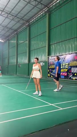 This media file features two people, a woman and a man, on a badminton court. The woman is wearing a white athletic outfit consisting of a sports bra and a pleated skirt, and white sneakers. She is holding a badminton racket and a shuttlecock. The man is wearing a blue t-shirt, black shorts, and white and blue sneakers. He is also holding a badminton racket.

They appear to be engaged in a badminton activity, possibly a practice session or a demonstration. The woman is shown in the process of serving or hitting the shuttlecock. The setting is an indoor badminton court with a green floor marked with white lines. The walls of the court are green, and the ceiling appears to be a metal structure.

In the background, a banner is visible with text that reads "ELITE Badminton CUP EP.3" and dates "6-7 DECEMBER 2025" and "8PM". This suggests that an Elite Badminton Cup event is being promoted or has taken place. The lighting indicates it is daytime, likely indoors. The overall atmosphere seems focused on the sport.

There are no specific cues about the weather, but the indoor setting implies it is not a factor. The location context provided is "บ้านบางรักษ์, Thailand," which helps to place the scene geographically. The emotions of the individuals are not explicitly clear from their postures, but their engagement with the sport suggests a concentration on the activity.
