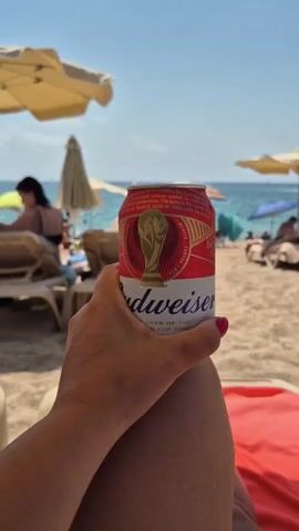 A close-up shot shows a hand holding a red and white can of Budweiser beer on a sunny beach. The can has a World Cup logo on it. The background is blurred, showing beach chairs, umbrellas, and the ocean. A person wearing a black swimsuit is visible in the distance, sitting on a beach chair.