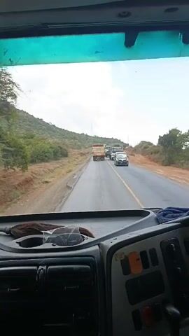 The video shows the view from the cab of a vehicle driving on a paved road. Three trucks are ahead of the vehicle, followed by a passenger van. Hills covered in trees and vegetation are visible on the left side of the road. The sky is cloudy. The dashboard of the vehicle is visible in the foreground, along with a piece of blue cloth.