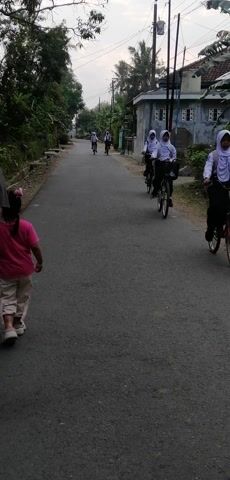 A video shows a group of people cycling and walking on a paved road in a rural area. The road is lined with trees and houses. In the foreground, a person in a pink shirt and beige pants is walking on the side of the road. Behind them, several children on bicycles are riding down the road. The sky is overcast. The video appears to be shot from a moving vehicle or by someone walking behind the group. The footage is recorded at a slightly tilted angle.