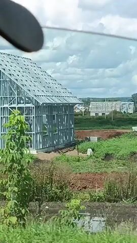 The video shows a developing residential area with several houses under construction. In the foreground, there are some plants and dirt. In the midground, there are several houses with metal framing and roofs, and some finished houses with gray walls and dark roofs. There is also construction equipment visible, including a yellow excavator. The background shows more houses, trees, and a cloudy sky. A black car is parked near one of the houses. The dialogue, which is in Russian, discusses whether bears are present in the area, with one person saying they only think so.